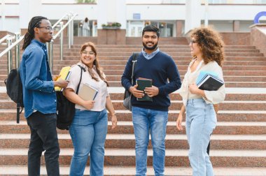 Üniversite kampüsünün merdivenlerinde duran, iletişim kuran, gülümseyen, kitap ve sırt çantalarını tutan bir grup genç öğrenci.
