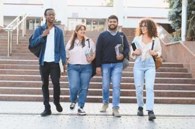 Bir grup çok ırklı üniversite öğrencisi kampüste birlikte yürüyor, ellerinde ders kitapları ve sırt çantaları, gülümsüyorlar.