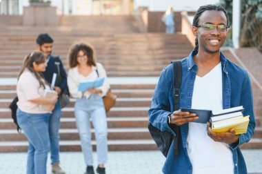 Elinde ders kitabı ve tablet olan gülümseyen erkek öğrenci, bir üniversite kampüsünde diğer çeşitli öğrencilerin önünde duruyor.