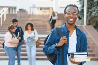 Sırtında kitap ve sırt çantası olan gülümseyen genç adam diğer öğrencilerle üniversite binasının önünde duruyor.