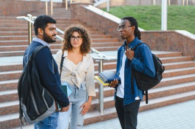 Bir üniversite kampüsünde granit merdivenlerde dururken, çeşitli öğrenciler ellerinde kitap ve sırt çantalarıyla tartışıyorlar.