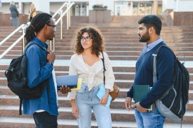 Çeşitli üniversite öğrencileri iletişim kuruyor, kitap ve sırt çantalarını tutuyor, kampüs merdivenlerinde duruyor, fikirlerini paylaşıyorlar.