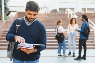 Üniversite kampüsünde kitap okuyan genç Hintli bir adam, arka planda çeşitli öğrencilerle konuşuyor.