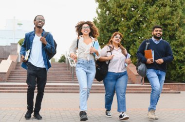 Group of happy young adult students carrying books and backpacks, walking down a path on a university campus