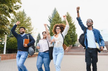 Group of diverse university students walking on campus, raising arms in celebration of achievement or graduation