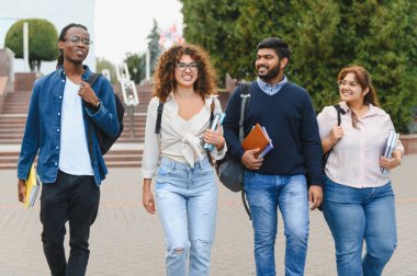 Üniversite kampüsünde kitap ve sırt çantaları taşıyarak birlikte yürüyen bir grup mutlu çok kültürlü öğrenci.