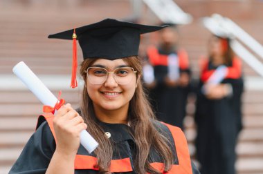 Young indian woman celebrating academic achievement, smiling and holding a rolled diploma. Graduates stand in the background