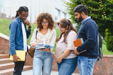 Group of diverse university students standing outdoors, reading books and discussing academic work during a break on campus