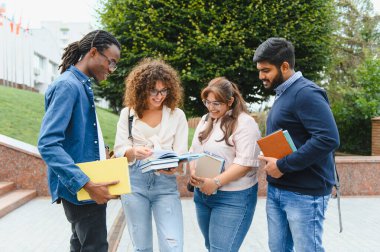 Group of four smiling diverse university students standing outdoors on campus, sharing textbooks and discussing studies