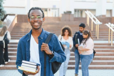 Confident young black man holding textbooks and backpack, smiling at camera, with diverse students walking on university steps