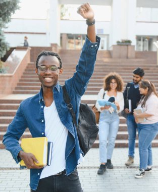 Smiling young man holding books, raising arm in celebration on campus with other diverse students in background