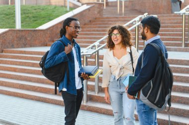 Farklı geçmişlere sahip üç genç öğrenci üniversite kampüsünün merdivenlerinde sırıtıyorlar, sırt çantaları ve kitaplar taşıyorlar.
