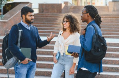 Çeşitli üniversite öğrencileri açık havada iletişim kuruyor, çantalar ve kitaplar taşıyor, arkadaşlık ve eğitim kavramlarını temsil ediyorlar.