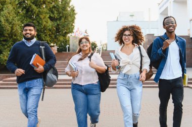 Diverse, happy young adult students walking on a university campus with books and backpacks, smiling and looking at camera