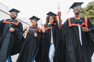 Group of smiling multi ethnic graduates wearing cap and gown, proudly showing diplomas after their university graduation ceremony