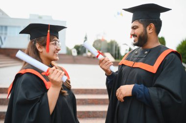 Two happy indian university students in graduation gowns and caps holding diplomas, celebrating their educational success