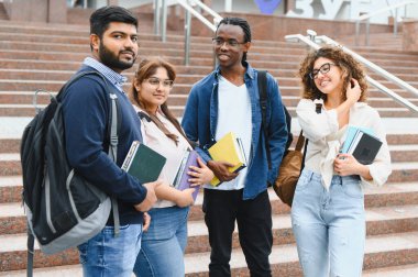 Diverse group of young students smiling, holding books while standing on university campus stairs, representing education and friendship