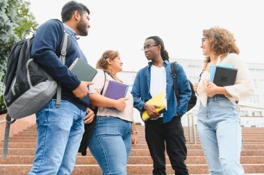 Group of multicultural college students standing on campus stairs, holding books and backpacks, discussing coursework