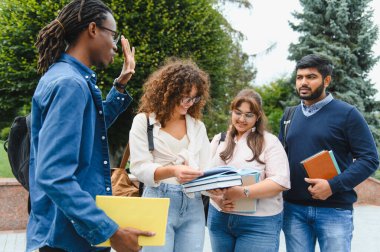 Group of multicultural university students discussing schoolwork, holding books and backpacks outdoors on campus
