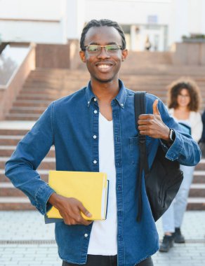 African american student smiling, holding books, giving a thumb up sign in front of a university building