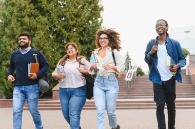 Group of happy young adults, diverse students, walking together on a university campus with backpacks and books