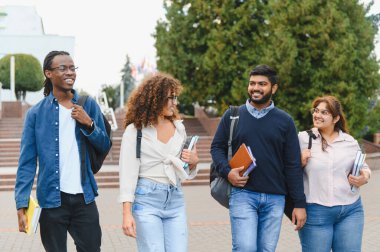 Group of multicultural young adults walking with books and backpacks outdoors, happy to be studying at college