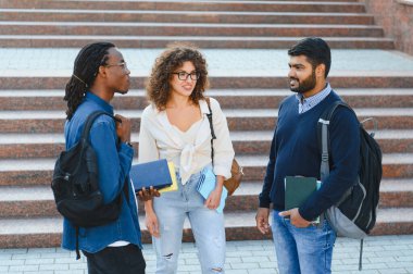 Three diverse students standing on campus stairs, discussing and smiling, holding books and wearing backpacks