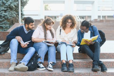 Diverse group of young university students sitting on outdoor stairs, reading books, collaborating, and discussing academic material