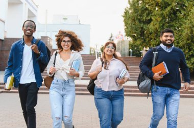 Happy multiethnic student group walking together, carrying books and backpacks, enjoying college life on campus
