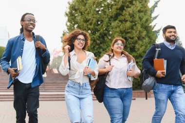 Group of happy multicultural university students carrying backpacks and books, walking together on campus