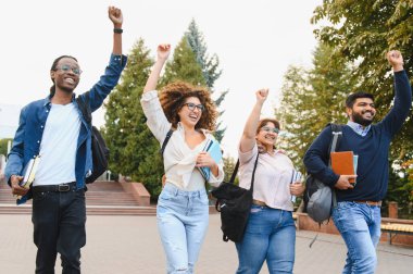 Group of happy diverse university students raising arms in celebration, walking on campus with books and backpacks