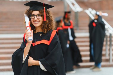 Young female graduate in gown and mortarboard proudly holding diploma on campus with smiling classmates celebrating success