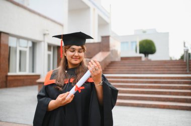 Indian woman student proudly holding her diploma while wearing cap and gown after completing her university education