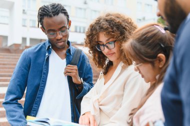 Group of multi ethnic students standing outdoors on university campus, attentively collaborating on a textbook