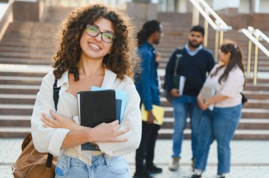 Smiling young woman with glasses and books standing on a university campus with diverse students in the background