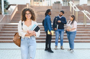 Smiling female student holding books with diverse friends chatting in the background, representing education and successful learning