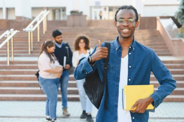 Young male student holding books and backpack, smiling and showing thumbs up on campus. Diverse student group in background