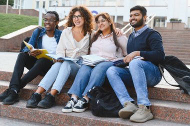 Group of happy multiracial university students sitting on outdoor steps, holding books and enjoying their diverse friendship
