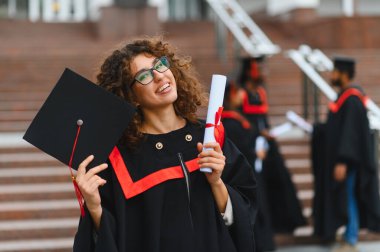 Happy female student celebrating graduation, wearing cap and gown, holding diploma with other graduates in background