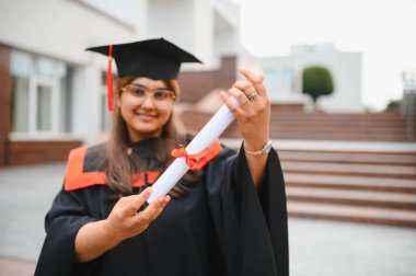 Indian graduate woman celebrating university achievement, holding her diploma scroll with joy, standing in academic dress