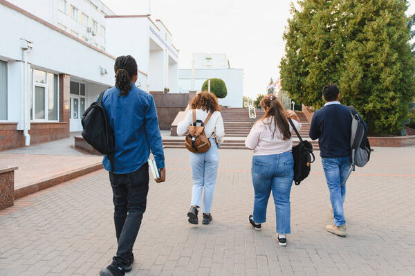 Diverse group of students walking along a paved path towards a university building, carrying backpacks and books