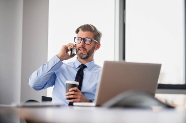 Yetişkin bir iş adamı, dizüstü bilgisayarla masada oturuyor ve cep telefonuyla konuşuyor. Ofiste çalışan iş adamı..