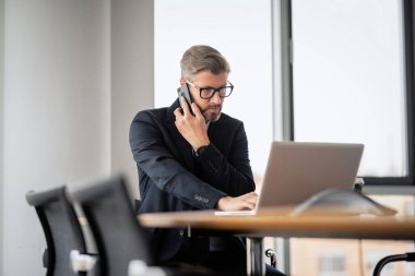 Yetişkin bir iş adamı, dizüstü bilgisayarla masada oturuyor ve cep telefonuyla konuşuyor. Ofiste çalışan iş adamı..