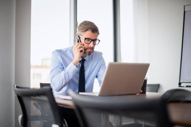 Profesyonel bir iş adamının dizüstü bilgisayarla masada oturup cep telefonuyla konuştuğunu düşünmek. İş adamı gömlek ve kravat takıyor..