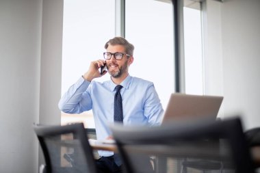 Yetişkin bir iş adamı, dizüstü bilgisayarla masada oturuyor ve cep telefonuyla konuşuyor. İşadamı ofiste çalışırken dizüstü bilgisayar kullanıyor..