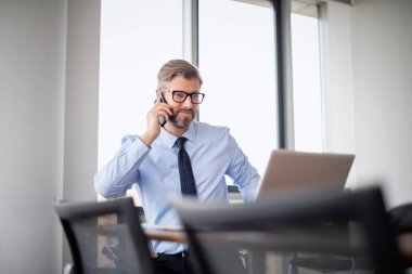 Yetişkin bir iş adamı, dizüstü bilgisayarla masada oturuyor ve cep telefonuyla konuşuyor. İşadamı ofiste çalışırken dizüstü bilgisayar kullanıyor..
