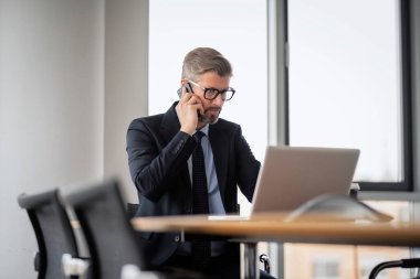 Yetişkin bir iş adamı, dizüstü bilgisayarla masada oturuyor ve cep telefonuyla konuşuyor. Ofiste çalışan iş adamı..