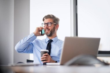 Yetişkin bir iş adamı, dizüstü bilgisayarla masada oturuyor ve cep telefonuyla konuşuyor. İşadamı ofiste çalışırken dizüstü bilgisayar kullanıyor..