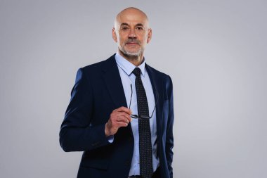 Gri arka planda duran kendine güvenen erkek CEO 'nun portresi. İş adamı takım elbise ve kravat takıyor ve elinde gözlük tutuyor. Boşluğu kopyala. 