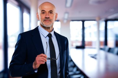 Ofiste duran kendinden emin erkek CEO 'nun portresi. Olgun yönetici takım elbise ve kravat takıyor. İş adamı kameraya bakarken elinde bir gözlük tutuyor..